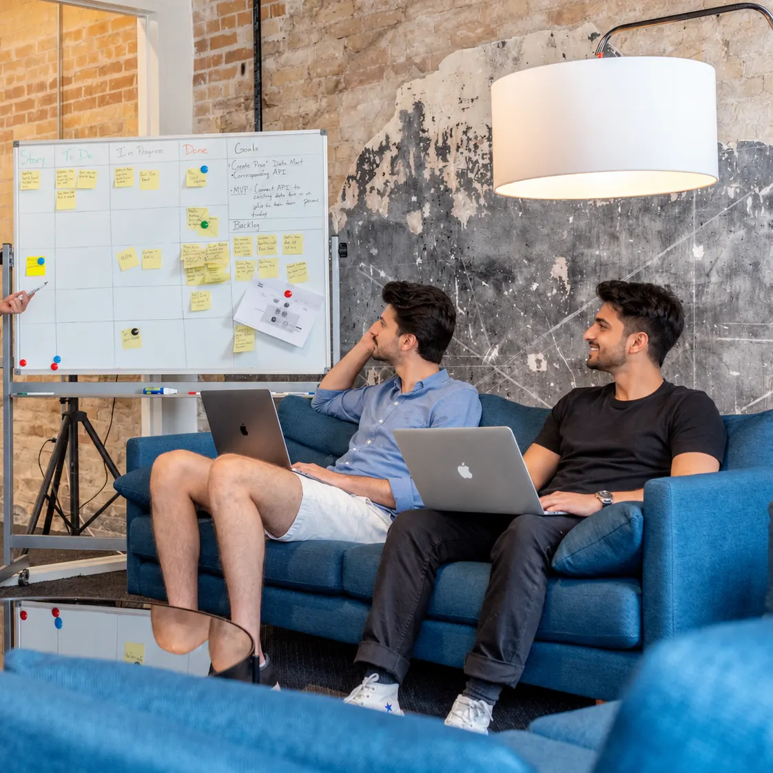 Two people on a sofa with laptops, planning a project beside a whiteboard of sticky notes